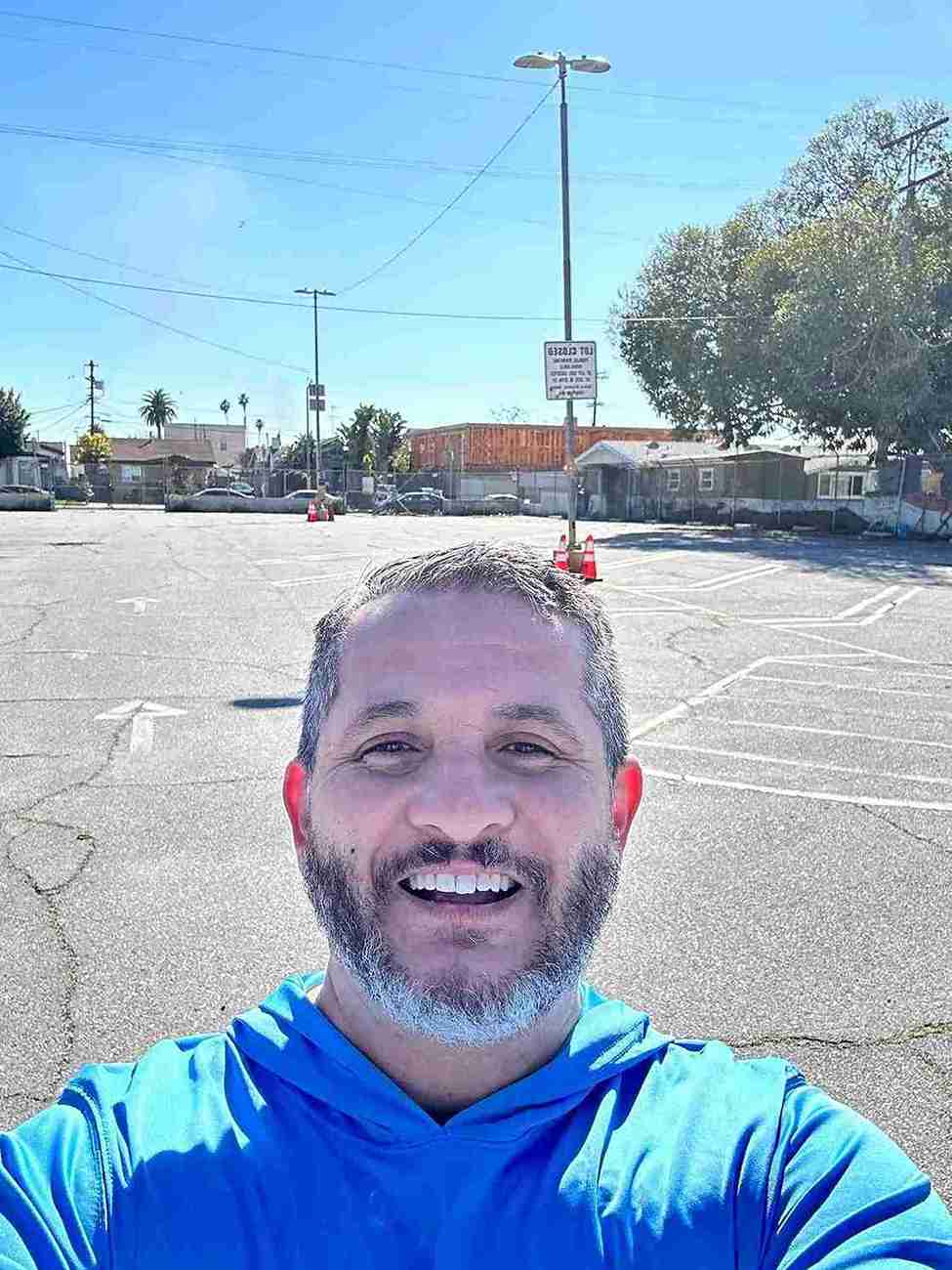 Man in a blue shirt standing in an empty parking lot with buildings and trees in the background.
